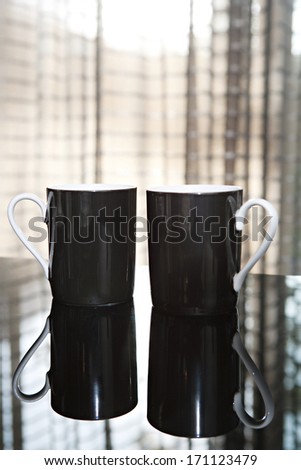 Still life close up detail view of two luxury black tea mugs on a reflective dining table in a quality expensive home with smart curtains. Home interior detail view.