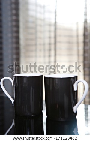 Still life close up detail view of a pair of luxury black tea mugs on a reflective dining table in a quality expensive home with smart curtains. Home interior detail view.