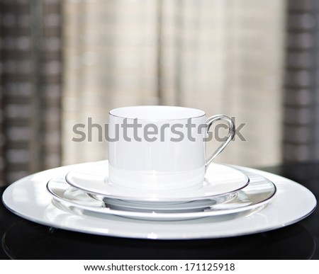 Close up still life detail view of a serving set of plates and cup for one on a luxury reflective diner table in a quality home with smart curtains. Home interior detail view.