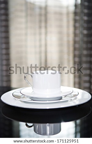 Close up still life detail view of a serving set of plates and cup for one on a luxury reflective diner table in a quality home with smart curtains. Home interior detail view.
