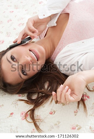Over head portrait view of an attractive young woman laying down on her bed in her home bedroom, having a telephone conversation on her smartphone and smiling. Home technology interior.