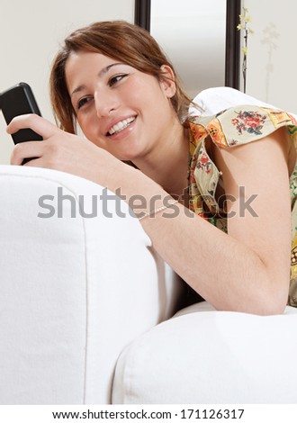 Attractive young woman lounging and relaxing on a white sofa in a home living room, holding and using a smartphone device while smiling. Home technology interior.