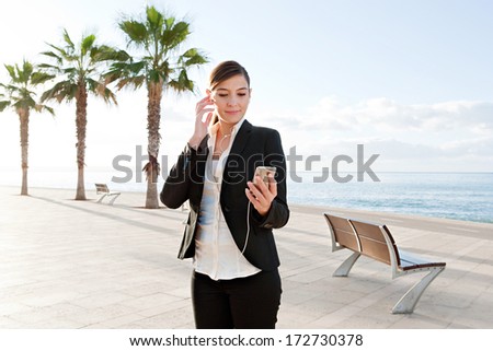 Portrait of a professional attractive businesswoman standing on a walk by the sea and listening to music using her smartphone and headphones. Business technology outdoors with a blue sky.