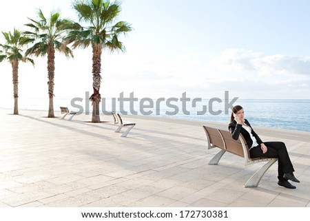Businesswoman sitting down on a bench on a walk by the sea, relaxing and having a mobile phone conversation using her smartphone during a sunny day at work. Business person outdoors.