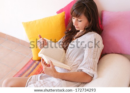 Close up portrait of a beautiful young woman on vacation, holding and reading a novel book while relaxing laying down on a colorful holiday sofa during a sunny summer day, interior.