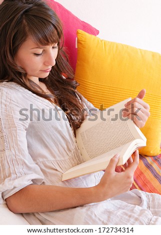 Close up portrait of a beautiful young woman on vacation, holding and reading a novel book while relaxing laying down on a colorful holiday sofa during a sunny summer day, interior.