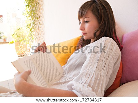 Close up portrait of a beautiful young woman on vacation, holding and reading a novel book while relaxing laying down on a colorful holiday sofa during a sunny summer day, exterior.