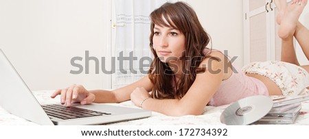 Panoramic portrait of a beautiful young woman relaxing on her bed at home using her laptop computer and typing, with films and music DVD\'s. Home entertainment interior lifestyle technology.