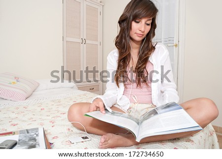 Attractive young woman sitting crossed legs on her bed at home and reading a magazine while listening to music with her headphones and mp3 player. Home entertainment technology interior.