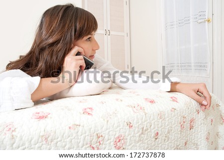 Close up profile portrait of an attractive young woman laying down on her bed relaxing at home and having a telephone conversation. Home interior lifestyle.