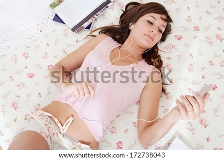 Over head portrait view of an attractive young woman laying down on her bed in her home bedroom using an mp4 player to listen to music with her headphones. Home technology interior.