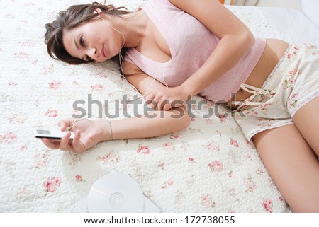 Over head portrait view of an attractive young woman laying down on her bed in her home bedroom using an mp4 player to listen to music with her headphones. Home technology interior.