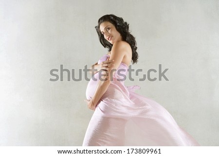 Side portrait of an attractive pregnant woman holding her belly with her hand while wearing a floating silk fabric dress against a plain background. Interior pregnancy beauty care.