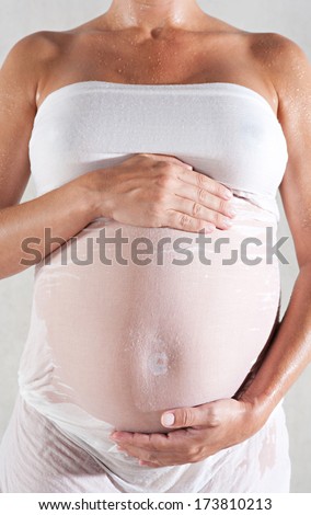 Close up faceless detail view of a pregnant woman body wearing a wet cotton fabric dress with her hands holding her belly. Interior pregnancy beauty care.