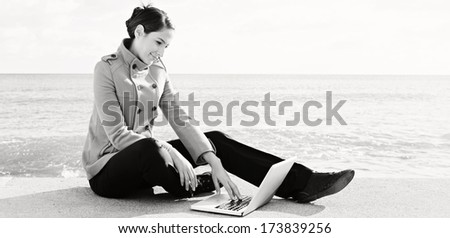 Black and white panoramic portrait of a young professional woman sitting down by the ocean with a sunny blue sky, using a laptop computer. Outdoors technology lifestyle.