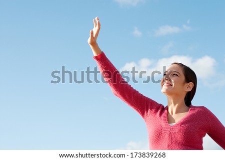 Portrait of an attractive young woman raising her hand to shade her face from the sun rays during a sunny day and against an intense blue sky, smiling. Beauty lifestyle exterior.