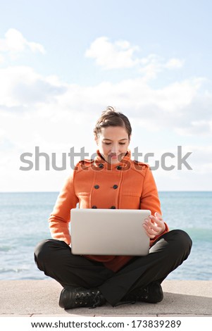 Frontal view of a young professional woman sitting down with her legs crossed by the ocean with a sunny blue sky, using a laptop computer and working on it during a break. Outdoors technology.