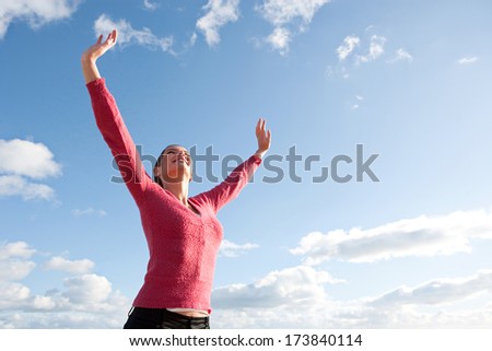 Joyful and attractive young woman feeling happy and raising her arms up to the intense blue sky in celebration. Fun and energetic expression on a colorful spacious sunny sky.