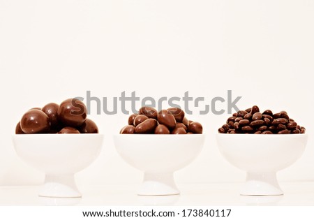 Close up detail view of a line of white porcelain containers holding different sizes of dark chocolate covered nuts on a white counter against a white background. Interior tempting food.