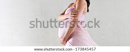Side view of a pregnant woman body figure wrapped in pink silk floating fabric, holding her belly with care against a spacious plain background. Interior beauty pregnancy portrait.