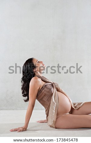 Profile view of an attractive pregnant woman sitting down and looking up while wrapped in transparent silk fabric against a spacious plain background. Interior pregnancy beauty.