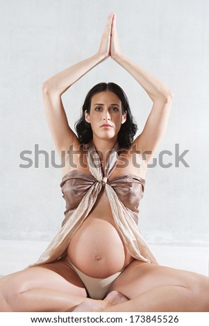 Close up of a pregnant woman sitting down in a yoga position, meditating and relaxing while wrapped in silk fabric against a plain background. Interior pregnancy health and beauty.