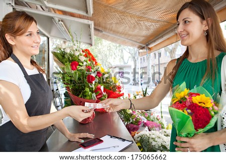 Attractive woman customer client playing with cash money for a small bouquet of sunflowers in a florist shop, with the business owner smiling happily and holding a bill.  Working business outdoors.