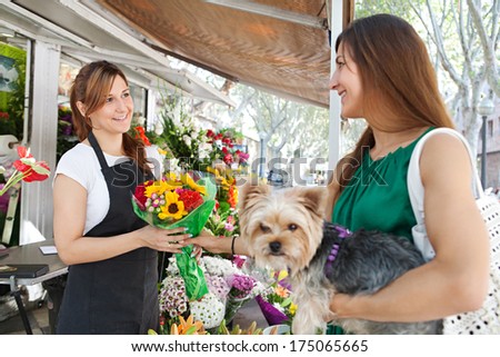 Florist business woman shop assistant selling a bouquet of fresh flowers to an attractive woman customer with her dog pet during a sunny day. Outdoors working business.