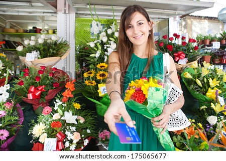 Portrait of an attractive florist store customer client woman buying a bouquet of fresh flowers and handing her credit card for payment in a flower market stall store. Outdoors business shopping.