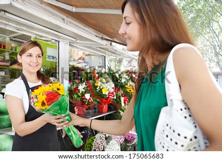Florist business woman shop assistant holding and selling a bouquet of fresh flowers to an attractive woman customer during a sunny day in a kiosk store. Outdoors working business.