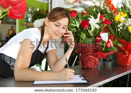 Close up portrait of a young and attractive florist business woman and shop attendant using a smartphone to take an oder over the phone during a sunny day. Working business outdoors.