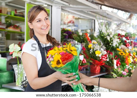 Close up portrait of a young and attractive florist business woman and shop attendant handing over a small bunch of sunflowers to her client during a sunny day. Working business outdoors.