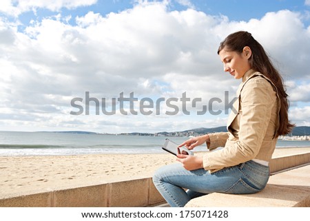 Side view of an attractive young woman sitting on a stone bench by the ocean sea using a digital tablet with touch screen during a sunny day out. Outdoors technology lifestyle.