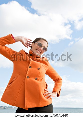 Attractive young woman being playful and posing with her orange coat being fun and quirky against a sunny blue sky with white clouds by the sea. Outdoors lifestyle coastal portrait.