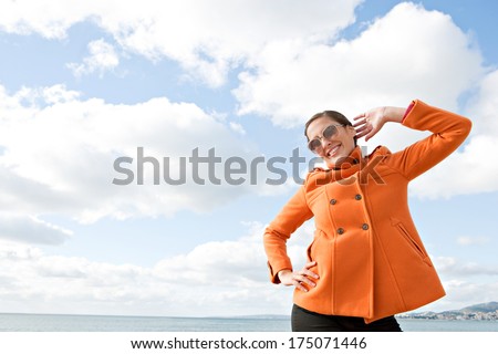 Attractive young woman being playful and posing with her orange coat being fun and quirky against a sunny blue sky with white clouds by the sea. Outdoors lifestyle coastal portrait.