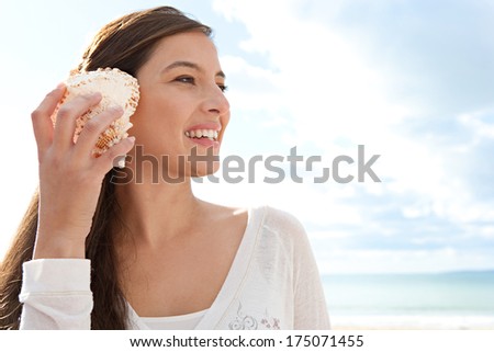Close up side portrait of a beautiful young woman on holiday holding a sea shell to her ear and smiling, listening to the sound of the ocean against a sunny blue sky. Outdoors lifestyle.