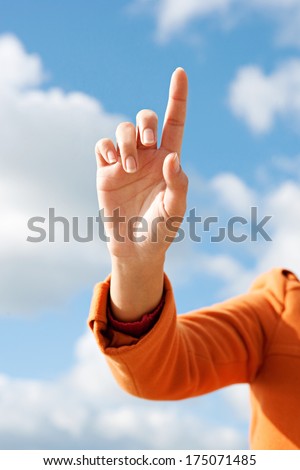Close up detail view of a woman hand and finger clicking or touching and imaginary button against a blue sky, wearing an orange coat during a sunny day, outdoors technology.