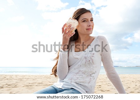 Portrait of a young attractive woman sitting on a white sand beach on vacation, holding a sea shell to her ear and listening to the ocean waves while relaxing and smiling. Outdoors holiday lifestyle.