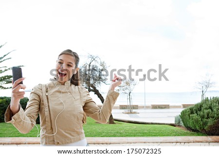 Fun And Attractive Young Woman Using Her Smartphone Device And Headphones To Sing, Dance And Listen To Music On Her Phone, Clicking Her Fingers Following The Rhythm. Outdoors Technology Lifestyle.