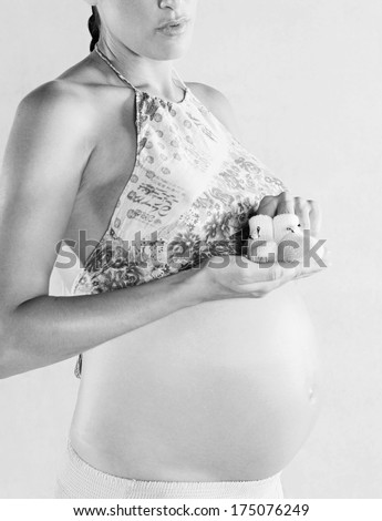 Black and white side body view of a pregnant woman showing her bare belly and holding a pair of crochet baby booties against a plain wall background. Pregnancy lifestyle, interior.