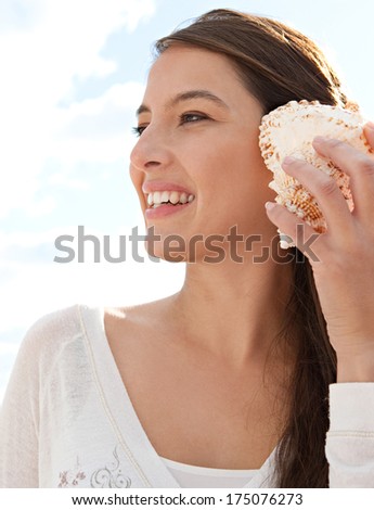 Close up side portrait of a beautiful young woman on holiday holding a sea shell to her ear and smiling, listening to the sound of the ocean against a sunny blue sky. Outdoors lifestyle.