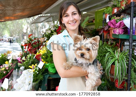 Portrait of an attractive florist kiosk business owner standing in her flower market stall shop, carrying her dog pet and smiling at the camera during a sunny day. Running a small business.