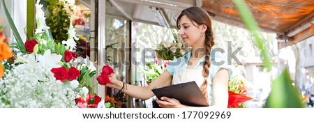 Panoramic side view of a florist small business owner checking her fresh flowers stock and inventory holding a clipboard, managing a market stall on a sunny day, outdoors. Running a small business.