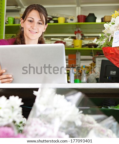 Portrait of an attractive florist business woman owner sitting at a flower shop counter using a laptop computer to place a stock order on line, smiling. Small business technology.