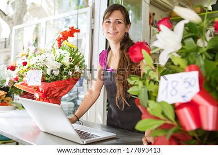 Portrait of a friendly and welcoming florist business woman owner proudly standing at the counter of a flower store using a laptop computer, smiling. Small business technology.