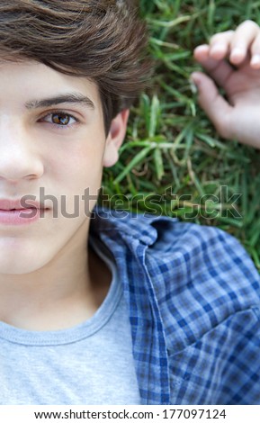 Over head close up portrait view of an attractive teenager student boy face laying down on healthy green grass in a park, relaxing and being thoughtful. Outdoors lifestyle.