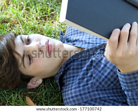 Over head portrait of an attractive young student boy holding up a text book from college and reading it, studying for exams while relaxing on green grass in a park. Outdoors lifestyle.