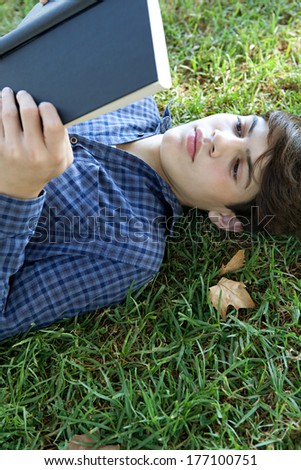 Over head portrait of an attractive young student boy holding up a text book from college and reading it, studying for exams while relaxing on green grass in a park. Outdoors lifestyle.
