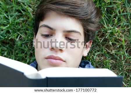 Over head close up portrait of an attractive young student boy holding up a text book from college and reading it, studying for exams while relaxing on green grass in a park. Outdoors lifestyle.