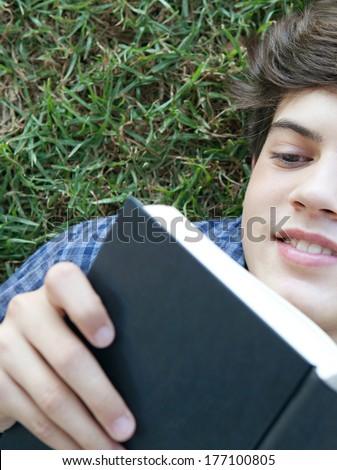 Over head close up portrait of an smiling young student boy holding up a text book from college and reading it, studying for exams while relaxing on green grass in a park. Outdoors lifestyle.
